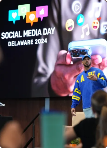 A speaker stands in front of a large screen displaying "SOCIAL MEDIA DAY DELAWARE 2024" and various social media icons.