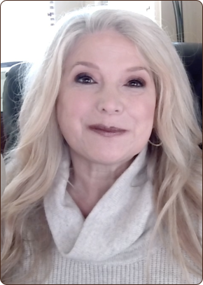 A blonde woman with long, wavy hair and makeup smiles, wearing a light-colored cowl-neck sweater.