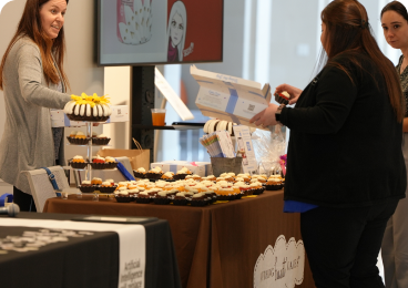 Two women stand behind a vendor table laden with small desserts.